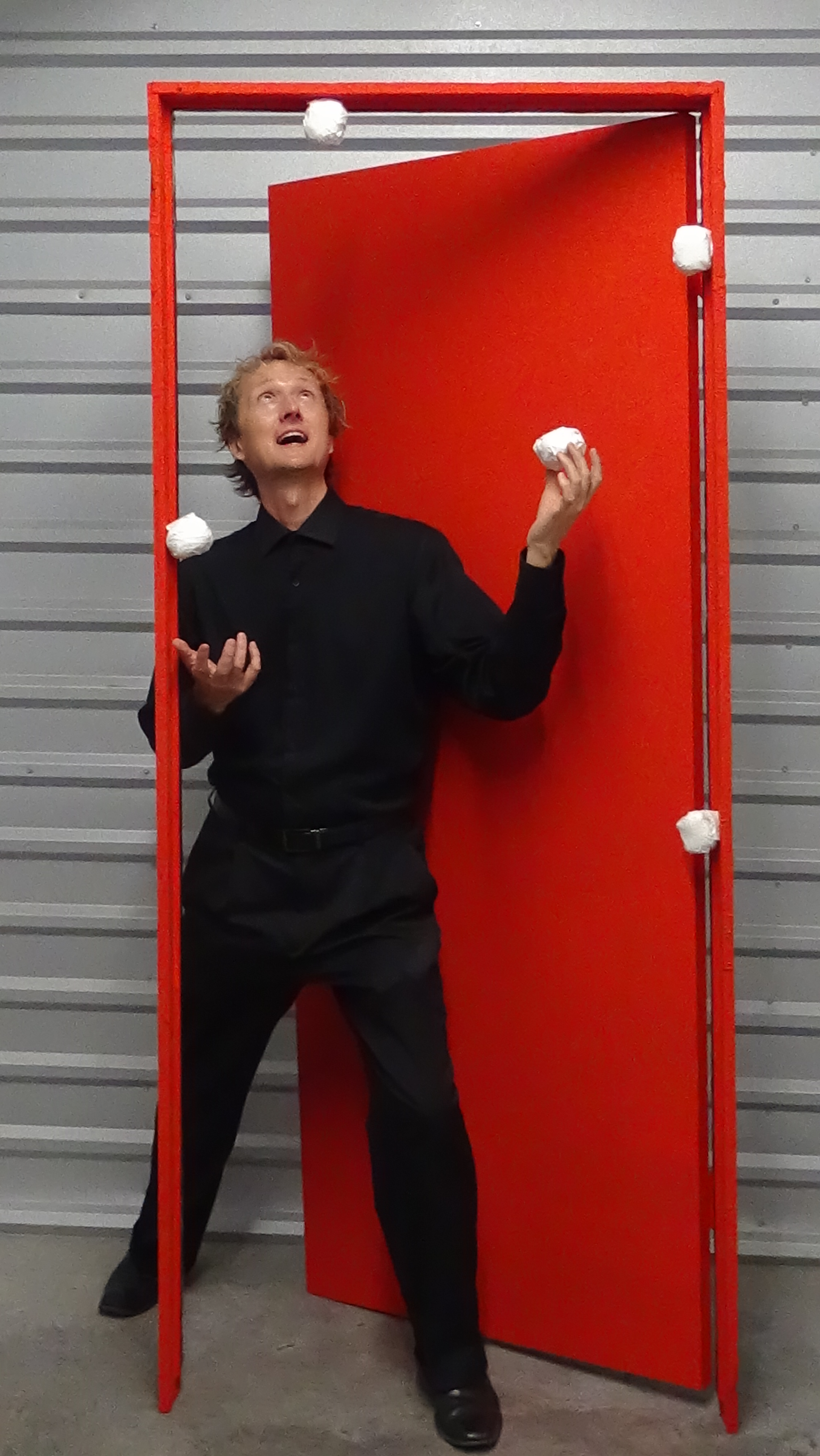 a juggler dressed in all black clothing stands against a bright red doorframe as he juggles four spherical objects