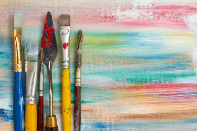 Image of a fine art pallet knife and paintbrushes of varying size and shape, on a wood plank background painted with horizontal streaks of different paint colors.