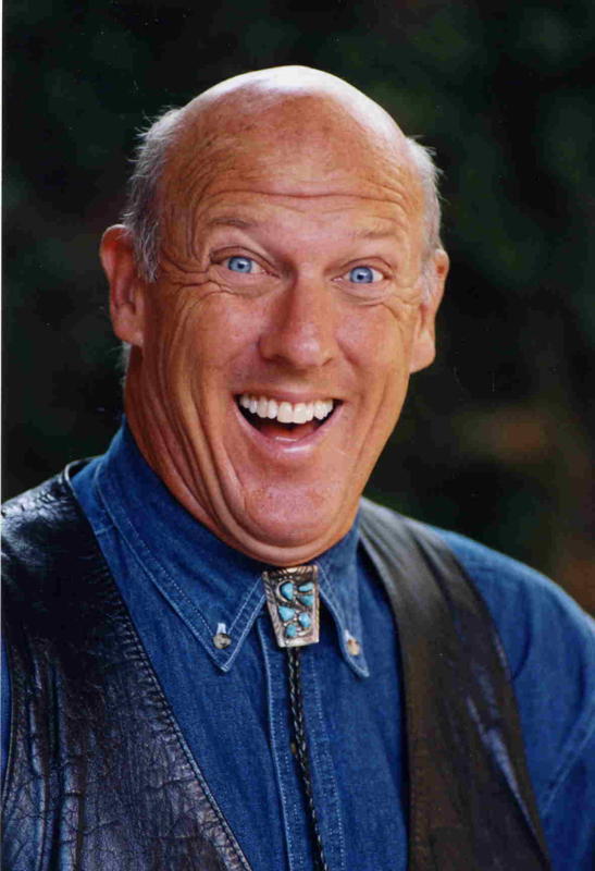 older bald man with blue eyes has an open, excited expression on his face. He is wearing an encrusted turquoise cowboy bolo tie, denim shirt, and black leather vest