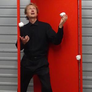 a juggler dressed in all black clothing stands against a bright red doorframe as he juggles four spherical objects