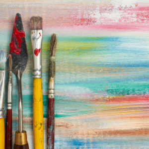 Image of a fine art pallet knife and paintbrushes of varying size and shape, on a wood plank background painted with horizontal streaks of different paint colors.